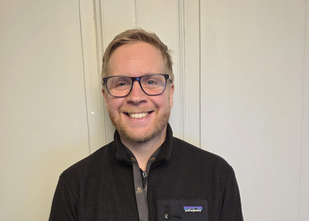 Porträttfoto av en person med kort ljust hår och glasögon som står framför en ljus, panelklädd vägg.