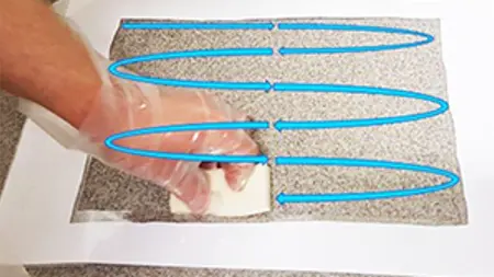 En hand för en trasa över ett golv. Illustrerade vågor som löper fram och tillbaka visar hur provtagningen gjordes. Foto.