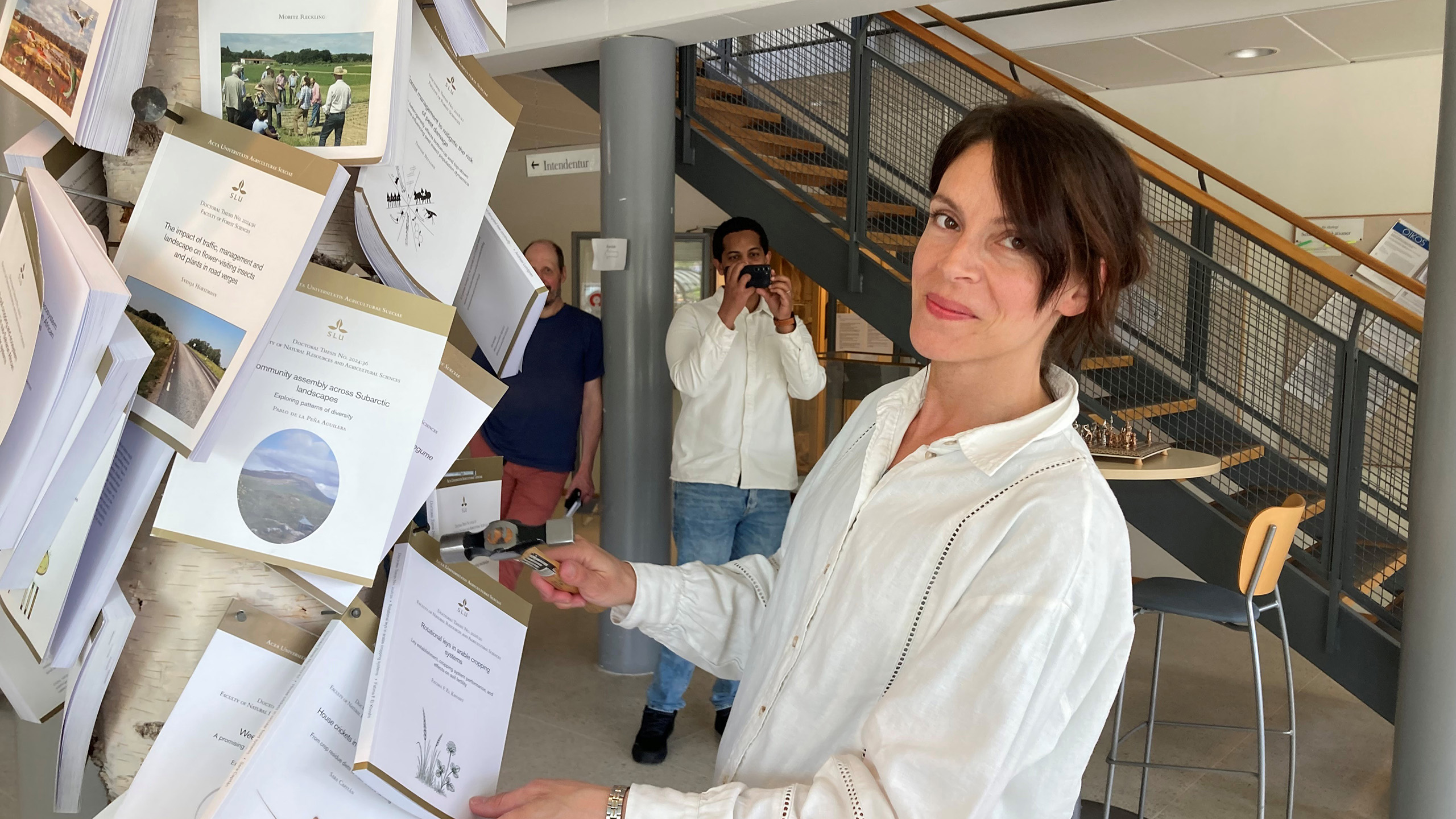 Fatima el Khosht håller i sin avhandling och en hammare. I bakgrunden står två personer, en håller sin mobilkamera.