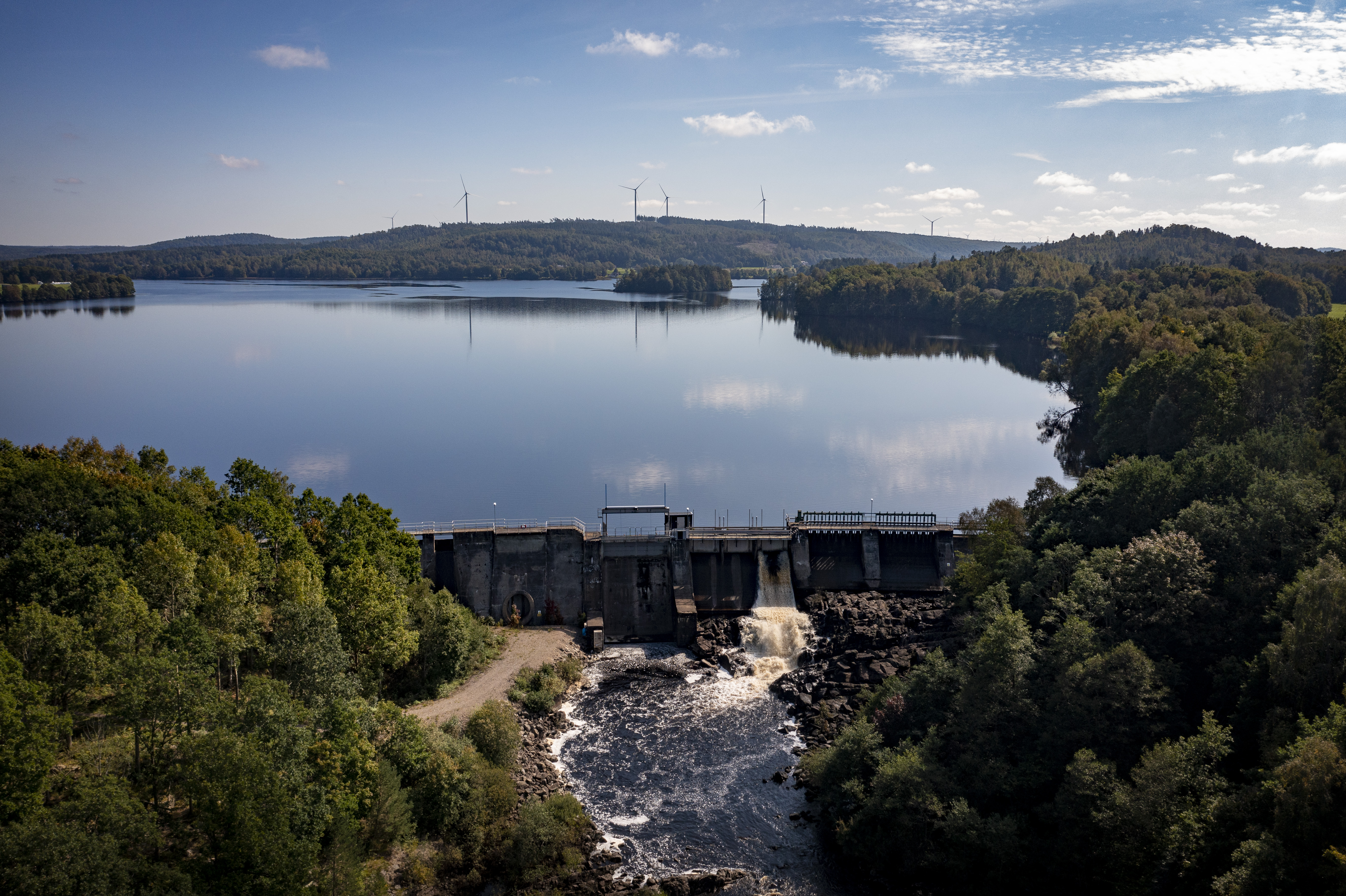 Vattenkraftsdamm med vatten som rinner nedströms, en större sjö uppströms och skogsklädda stränder runt omkring.