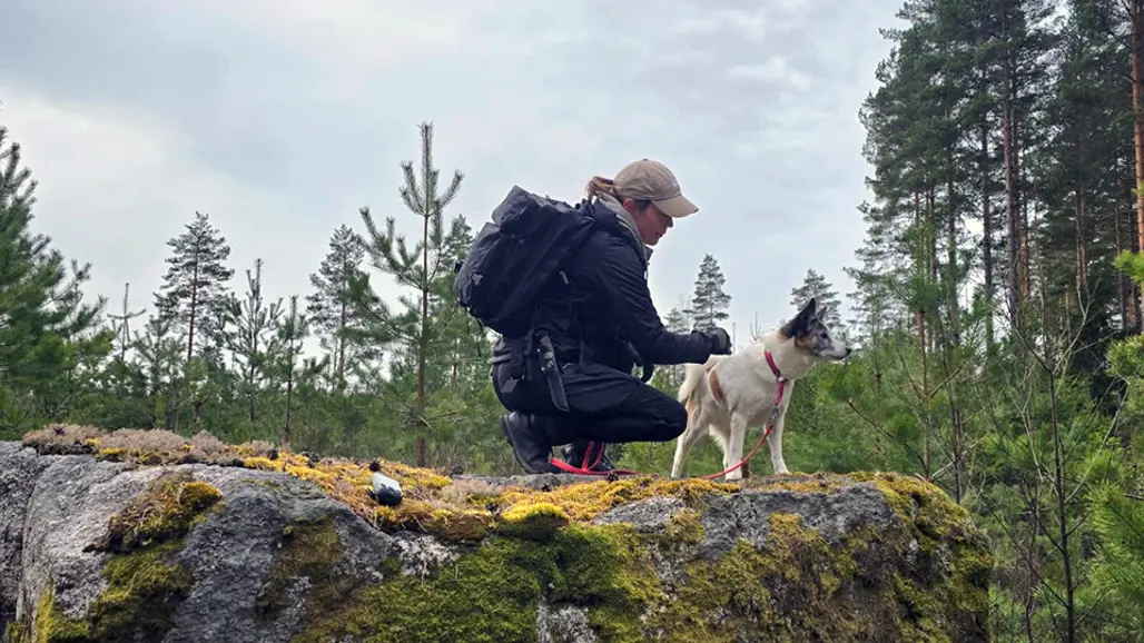 En person med ryggsäck och keps knäböjer på en stor mossbeklädd sten i skogen och klappar en liten hund som står bredvid. I bakgrunden syns höga träd och en mulen himmel. Foto.