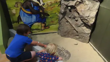 Två barn sitter framför en monter. I montern finns en grävning och en sten.