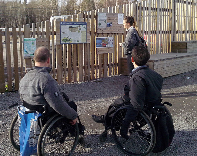 Tre personer står framför en informationsskylt. Två av personerna sitter i rullstol.