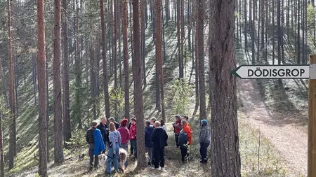 En grupp står och tittar på en dödisgrop. Gles tallskog omger dem. Det är kuperat där de står på en sandås.