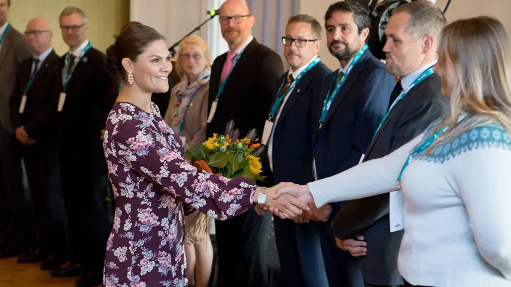 En person i en lila klänning med blommönster skakar hand med en person i en vit tröja med mönstrat band. Personen i klänningen håller en bukett med blommor. Bakom dem står flera personer i formella kläder med namnskyltar, i en inomhusmiljö med neutral bakgrund.