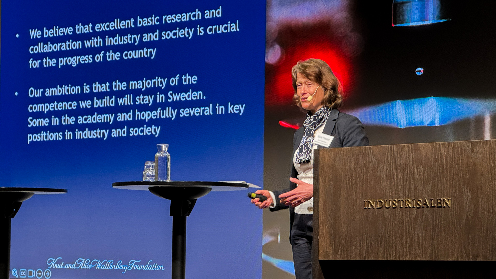 Person presenting on stage at a conference, with a slide projected in the background.