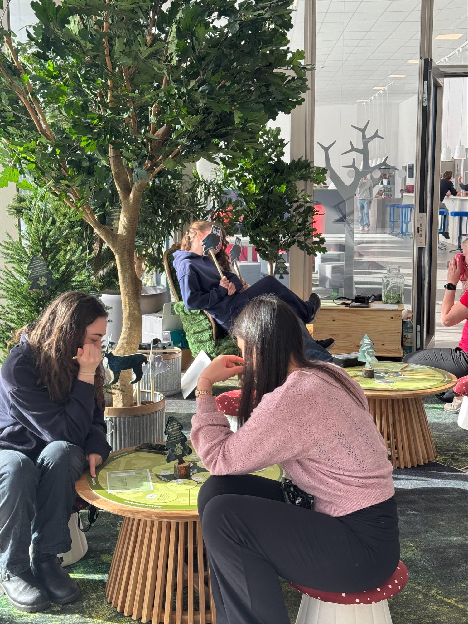 Personer sitter runt små runda bord i en ljus inomhusmiljö med grönska och trädliknande växter. De spelar ett brädspel tillsammans i en avslappnad miljö med soffor, stolar och glasväggar i bakgrunden. Foto.