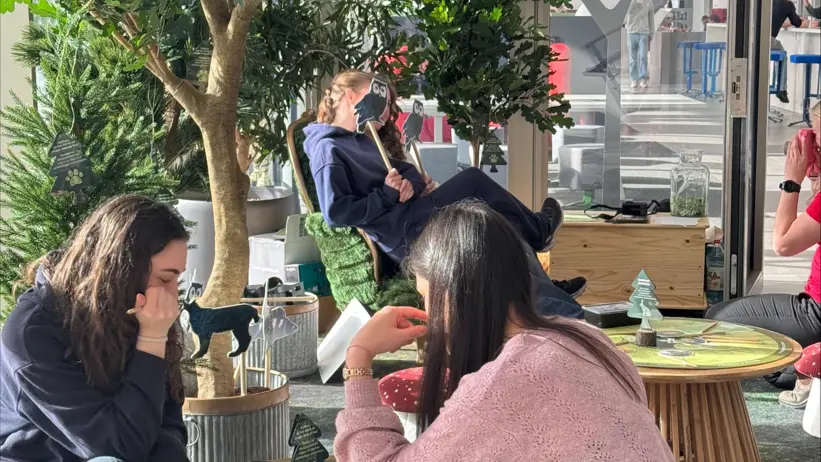 Personer sitter runt små runda bord i en ljus inomhusmiljö med grönska och trädliknande växter. De spelar ett brädspel tillsammans i en avslappnad miljö med soffor, stolar och glasväggar i bakgrunden. Foto.