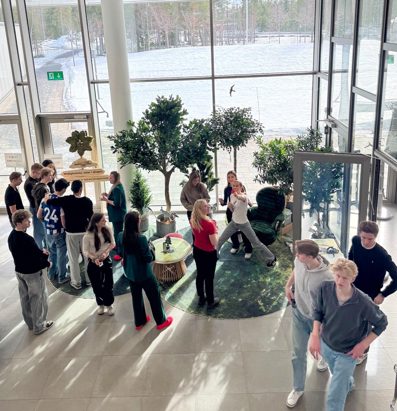 En grupp människor samlas i en ljus och rymlig inomhusmiljö med stora fönster mot ett snötäckt landskap. De står och rör sig runt en cirkulär sittgrupp med små bord och krukväxter, där samtal och aktiviteter pågår i en öppen och social atmosfär. Foto.