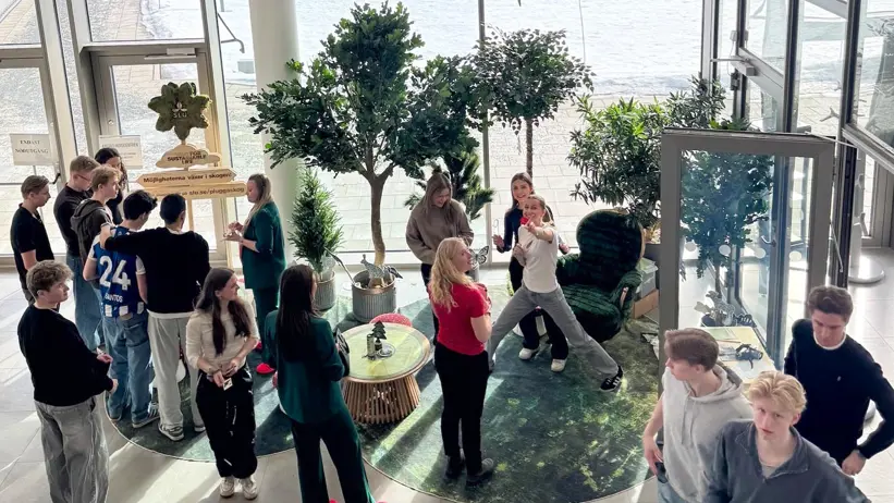 En grupp människor samlas i en ljus och rymlig inomhusmiljö med stora fönster mot ett snötäckt landskap. De står och rör sig runt en cirkulär sittgrupp med små bord och krukväxter, där samtal och aktiviteter pågår i en öppen och social atmosfär. Foto.