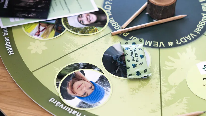 Närbild av ett runt brädspel på ett bord med grönt spelbräde, cirkelformade bilder, träpinnar och spelmarkörer. På spelbrädet ligger även en inslagen fiberbar och informationsmaterial, vilket ger intryck av en pågående spelaktivitet. Foto.