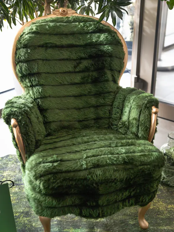 En grön, mjuk fåtölj med tjock, pälsliknande klädsel och trädetaljer står i en ljus inomhusmiljö. Runt stolen finns växter och ett fönsterparti som släpper in dagsljus och förstärker det naturinspirerade uttrycket. Foto.