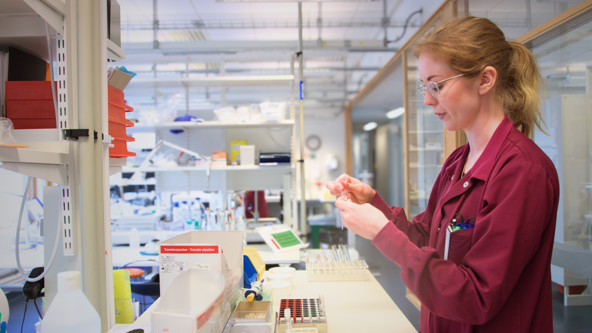 Woman with blond hair, glasses and wearing a red coat standing up. In front of her is a table with lab equipment. In the background are shelves with objects.