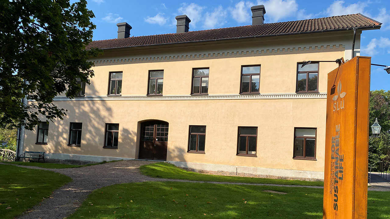 Sandstensfärgad byggnad omgiven av gröna träd