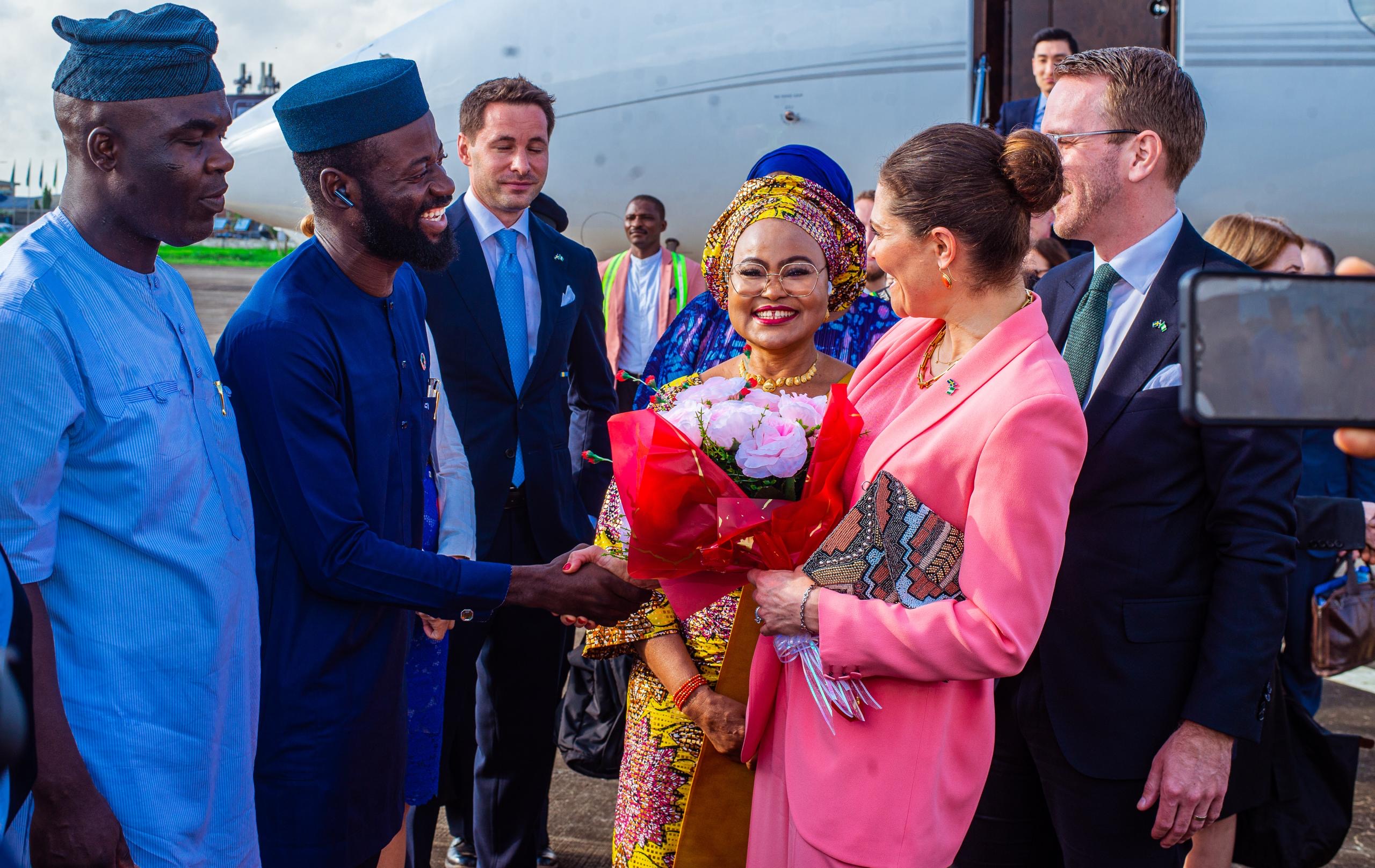 Delegation greeting beside an aircraft; a woman in pink receives flowers and shakes hands with a man in blue traditional attire.