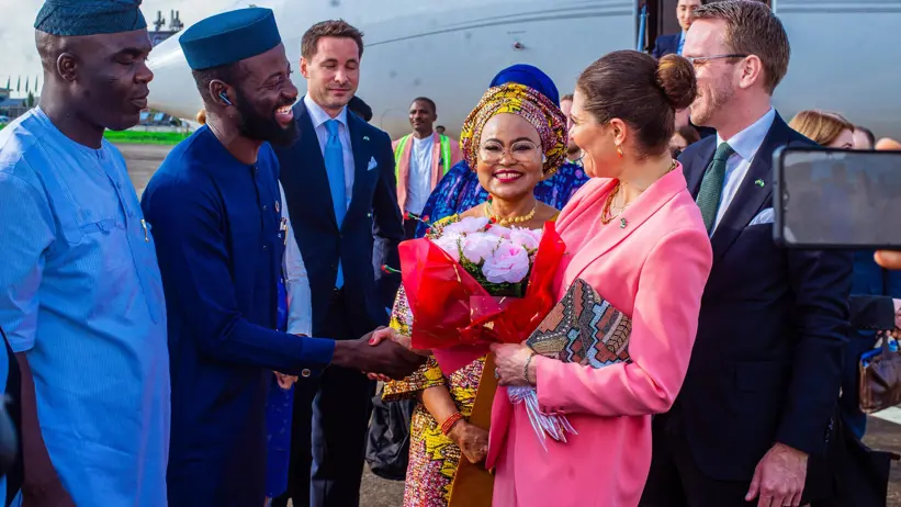 Delegation greeting beside an aircraft; a woman in pink receives flowers and shakes hands with a man in blue traditional attire.