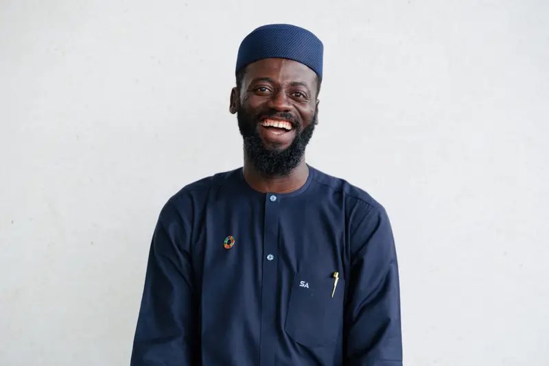 Smiling man in dark blue traditional clothing and cap against a plain background.