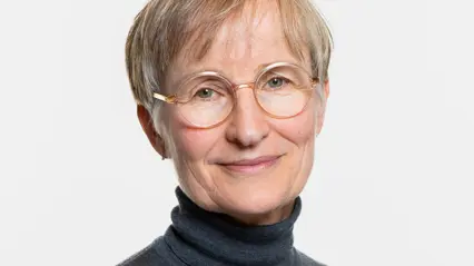 En person med kort, ljust hår och runda glasögon står mot en neutral bakgrund. Personen bär en mörk, högkragad tröja och ler svagt.