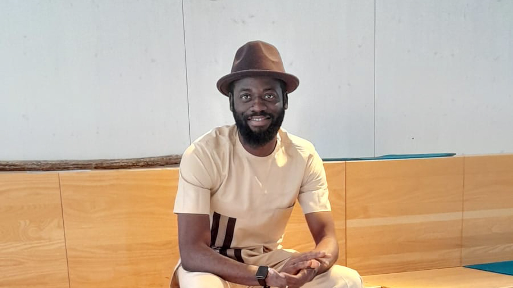 A smiling bearded person wearing a brown hat and a light beige outfit with dark details sits casually on a wooden bench. The background features a wood-panel and concrete wall.
