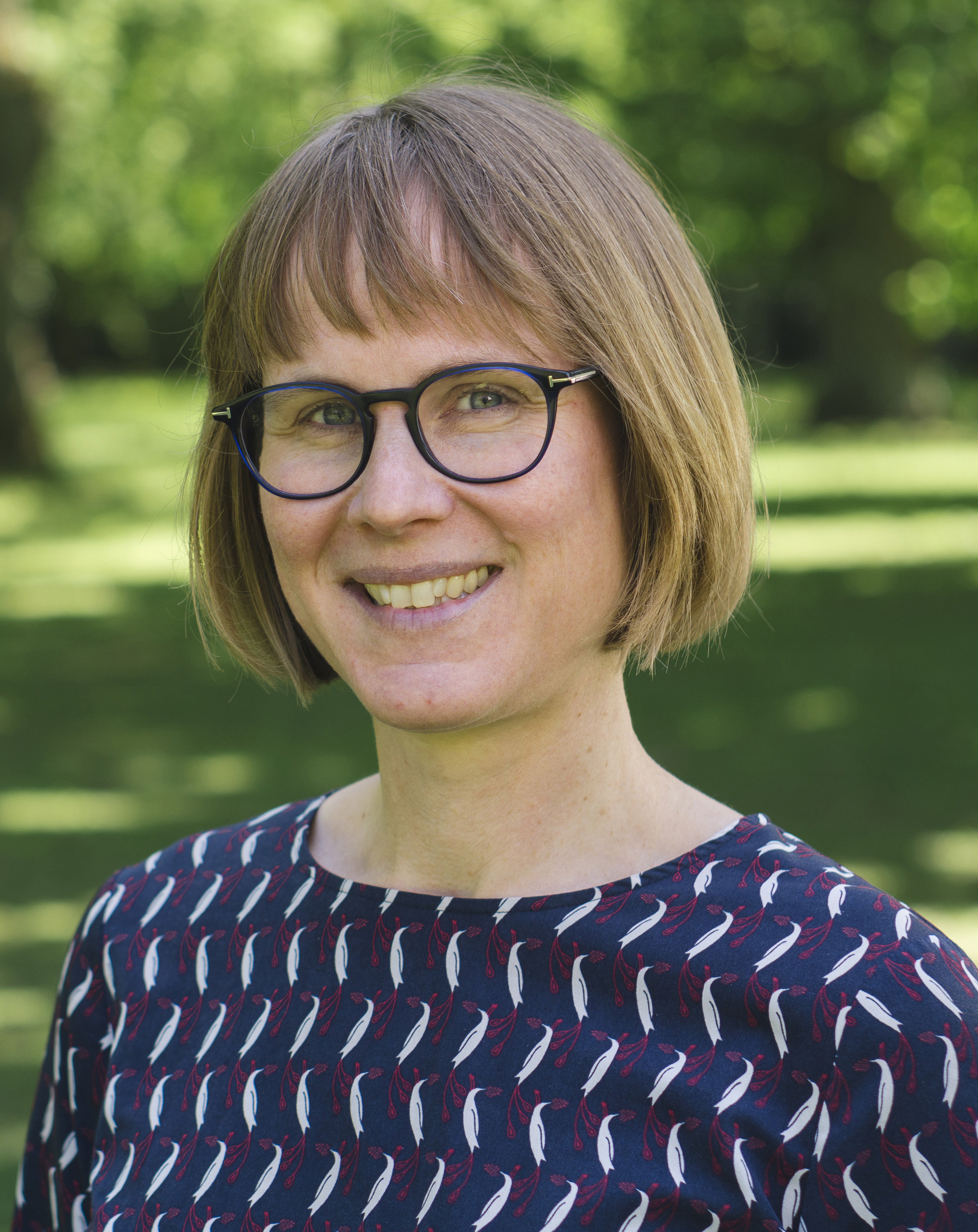 Portrait of a smiling woman with glasses and short hair, outdoors in front of a green park.