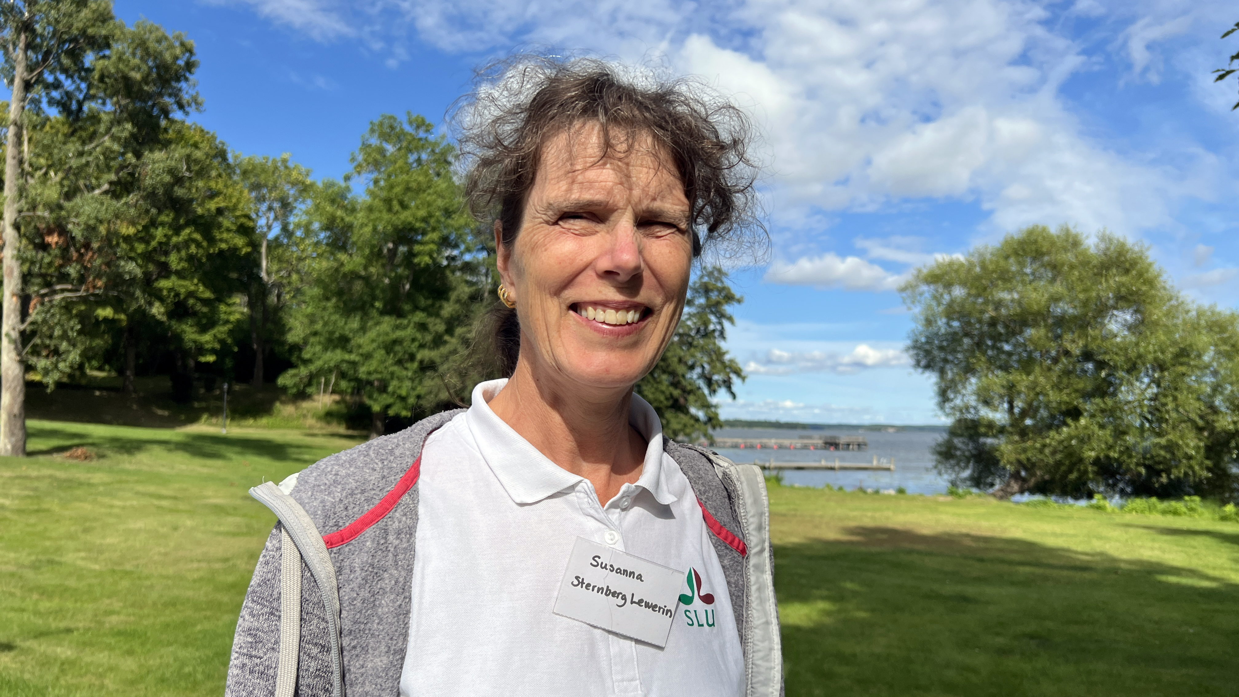 En kvinna som ler. I bakgrunden syns grönska och vatten. Foto.