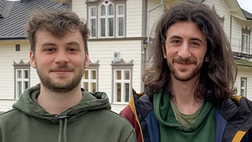 Två unga män med mörkt hår står framför en ljus träbyggnad med flera fönster och mörkt tak. Den ena mannen bär en grön huvtröja, och den andra har en grön tröja under en jacka. Foto.