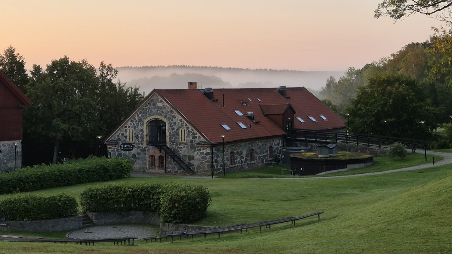 Stenbyggnad med rött tak i grön parkmiljö.