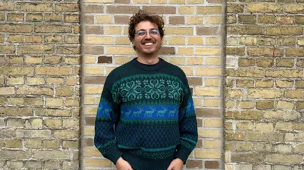 A smiling man stands in front of a light brick wall, wearing a dark green and blue patterned sweater with hands in his pockets.