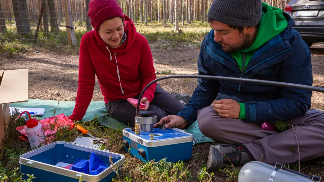Two people sitting on the ground in the forest, working with an instrument in a blue box