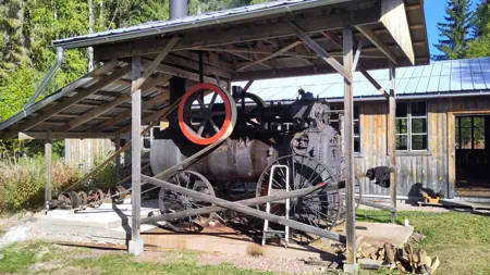 A large old steam driven machine under a roof in front of a wooden building.