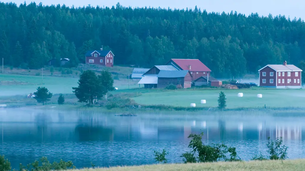 Ett norrländskt landskap med en älv, skog och röda hus. Foto.
