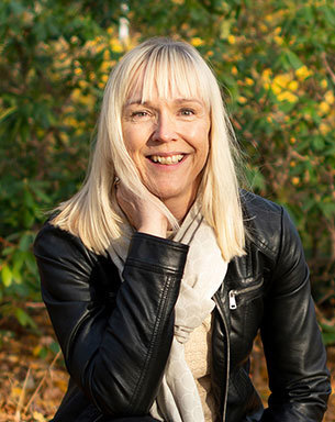 A woman with shoulder-length blonde hair and a black leather jacket is crouching among autumn leaves.