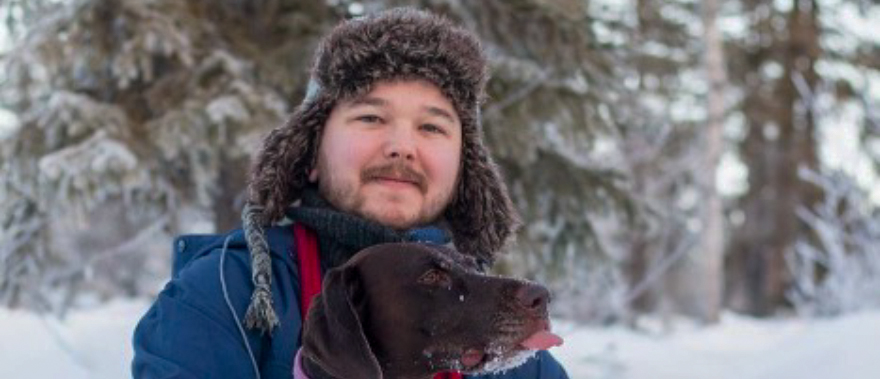 En person står utomhus i ett snötäckt landskap, iklädd en pälsfodrad vintermössa, halsduk och blå jacka. Personen håller en mörkbrun hund med röd halsband i famnen. Bakgrunden består av snötäckta träd som ger en lugn vinterkänsla. Foto.