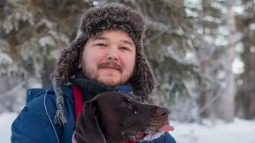 En person står utomhus i ett snötäckt landskap, iklädd en pälsfodrad vintermössa, halsduk och blå jacka. Personen håller en mörkbrun hund med röd halsband i famnen. Bakgrunden består av snötäckta träd som ger en lugn vinterkänsla. Foto.