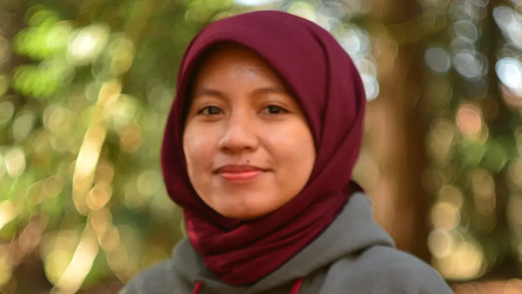 A person wearing a grey hoodie smiles at the camera. The background is softly blurred with green and golden light.