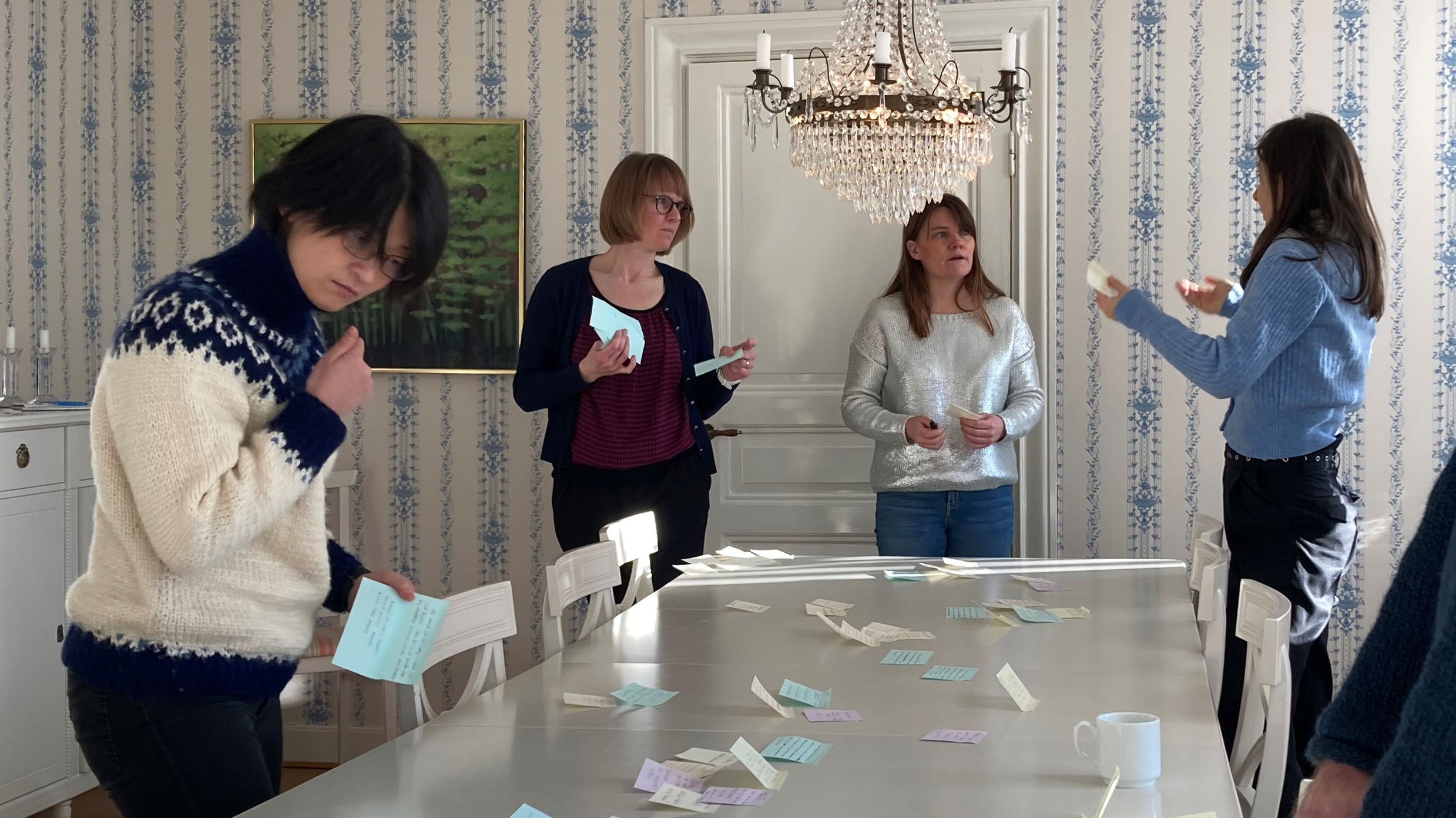 Fyra kvinnor står runt ett vitt bord under en kristallkrona och sorterar post-it-lappar. Foto.