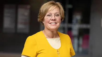 En person med kort, ljust hår och glasögon står i en inomhusmiljö med oskarp bakgrund. Personen bär en gul tröja över en beige topp och ler mot kameran.