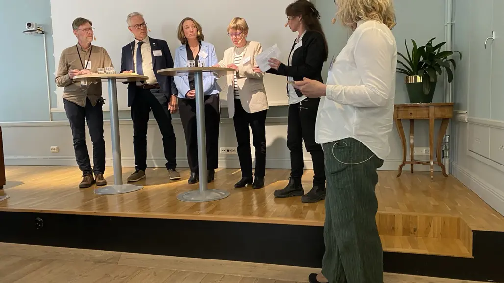 En grupp människor står uppradade och diskuterar vid några ståbord. Foto.