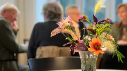 En vas med blommor i förgrunden och samtal mellan fyra personer i bakgrunden