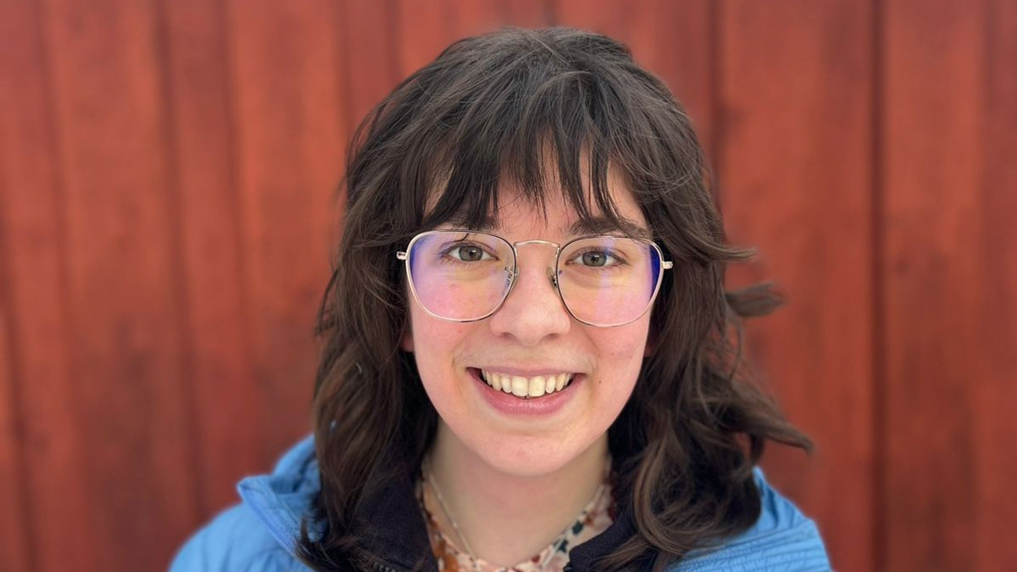 A person with wavy dark hair and glasses smiles at the camera. They are wearing a blue jacket and standing in front of a red wooden wall in daylight.