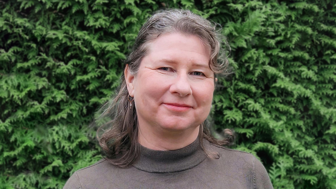 Kvinna med mörkgrått, lockigt hår och grå polotröja står framför grön häck. Foto