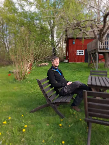 Man sitter på en trästol i en lummig trädgård framför ett rött hus och håller en kopp, omgiven av gräs, träd och vårblommor.