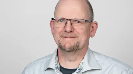 En person med kort, ljust hår och glasögon står mot en neutral bakgrund. Personen bär en ljus skjorta.