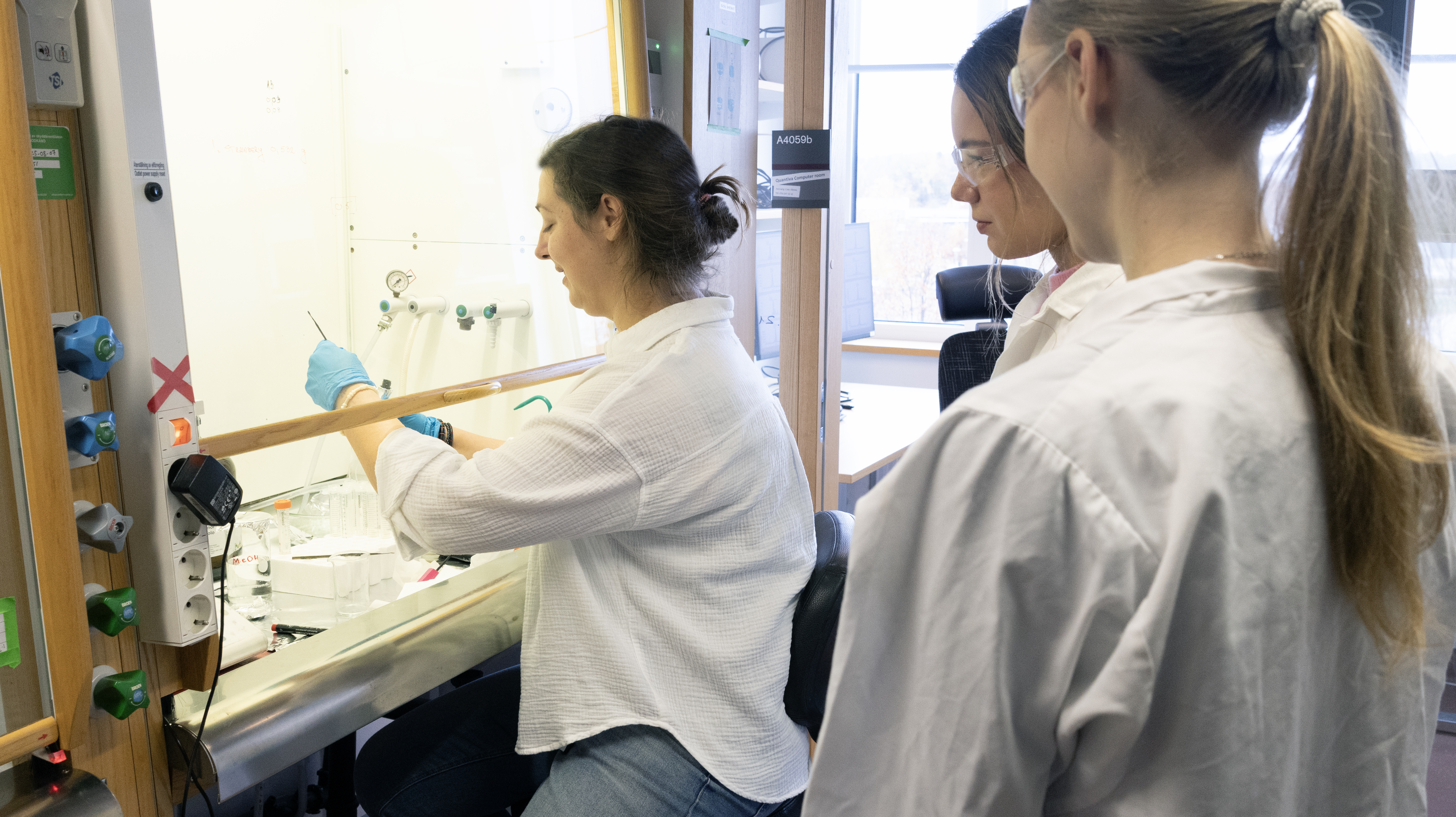 Elever flankerar labbföreståndare som arbetar vid dragskåp. Foto.