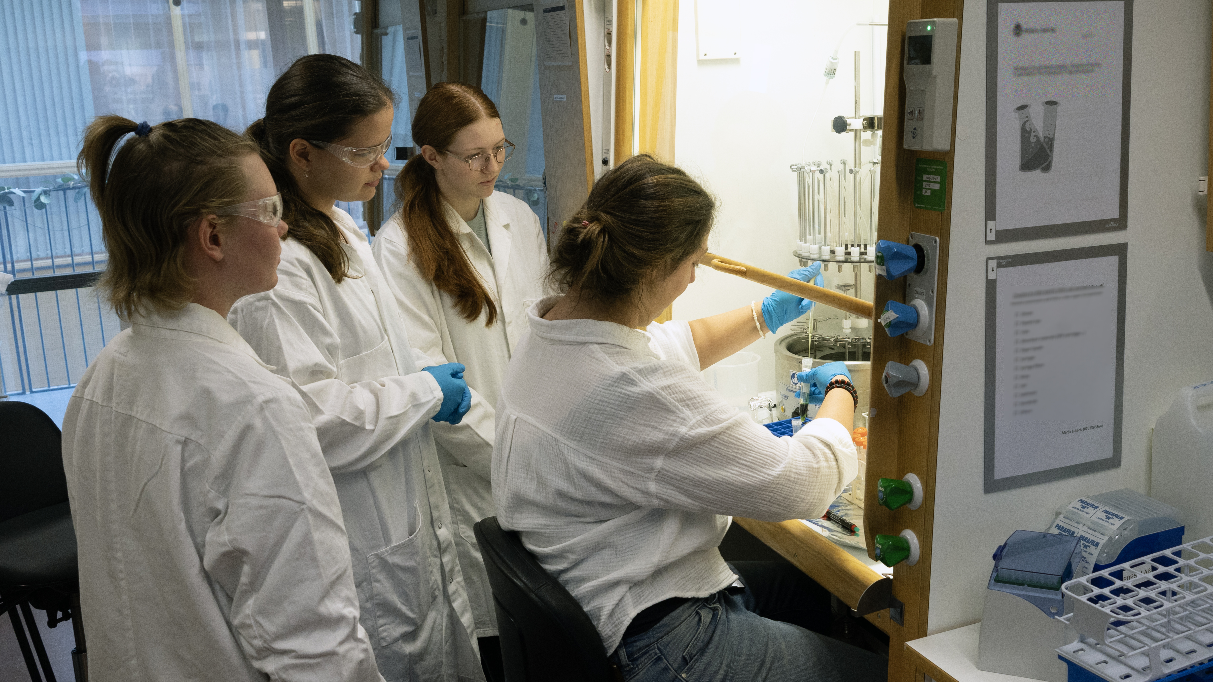 Elever flankerar labbföreståndare som arbetar vid dragskåp. Foto.