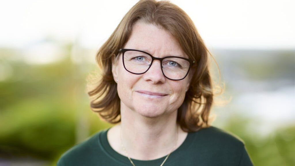 Brunhårig kvinna med glasögon och grön tröja framför suddig miljö med grönska och vatten. Foto