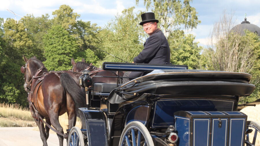 Anders Eriksson som finklädd och med hatt kör en mörkblå hästvagn med två mörkbruna hästar framför.