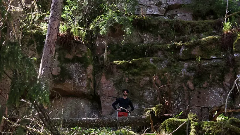 En person står i ett skogsområde med tät grönska och mossbeklädda klippor. Personen bär mörka kläder och verkar titta mot kameran. I bakgrunden syns höga träd och en klippvägg täckt av mossa och växtlighet. Foto.