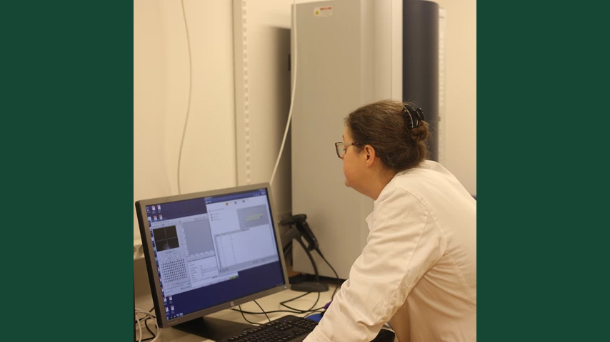 Photo of a woman in a lab coat in front of a computer-driven machine indoors. 