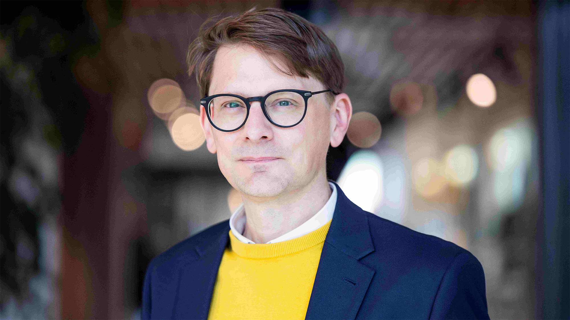 Brunhårig, slätrakad man med runda glasögon, marinblå kavaj, gul tröja och vit skjorta. Foto